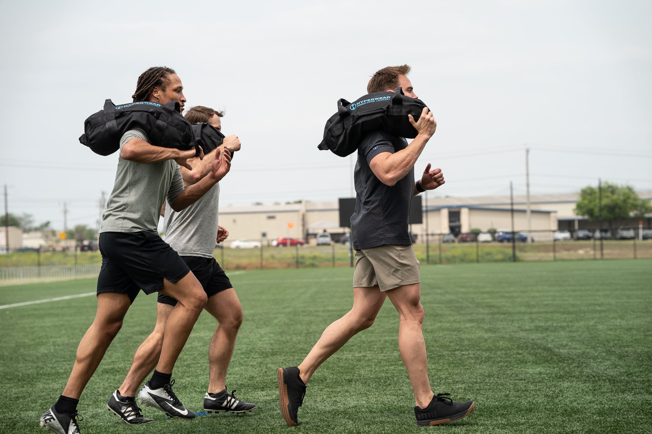 picture of 3 male athlete models on green field shouldering workout sandbags while running