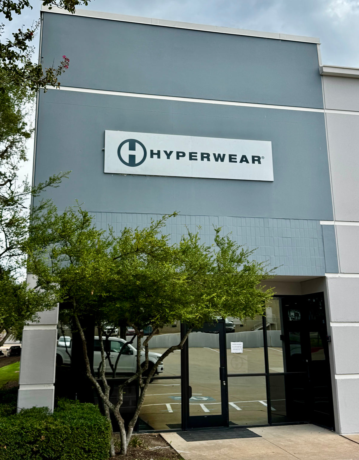 The exterior of a Hyperwear retail store building, featuring a gray facade, a white sign with the Hyperwear logo, and a glass entrance door partially obscured by green trees.
