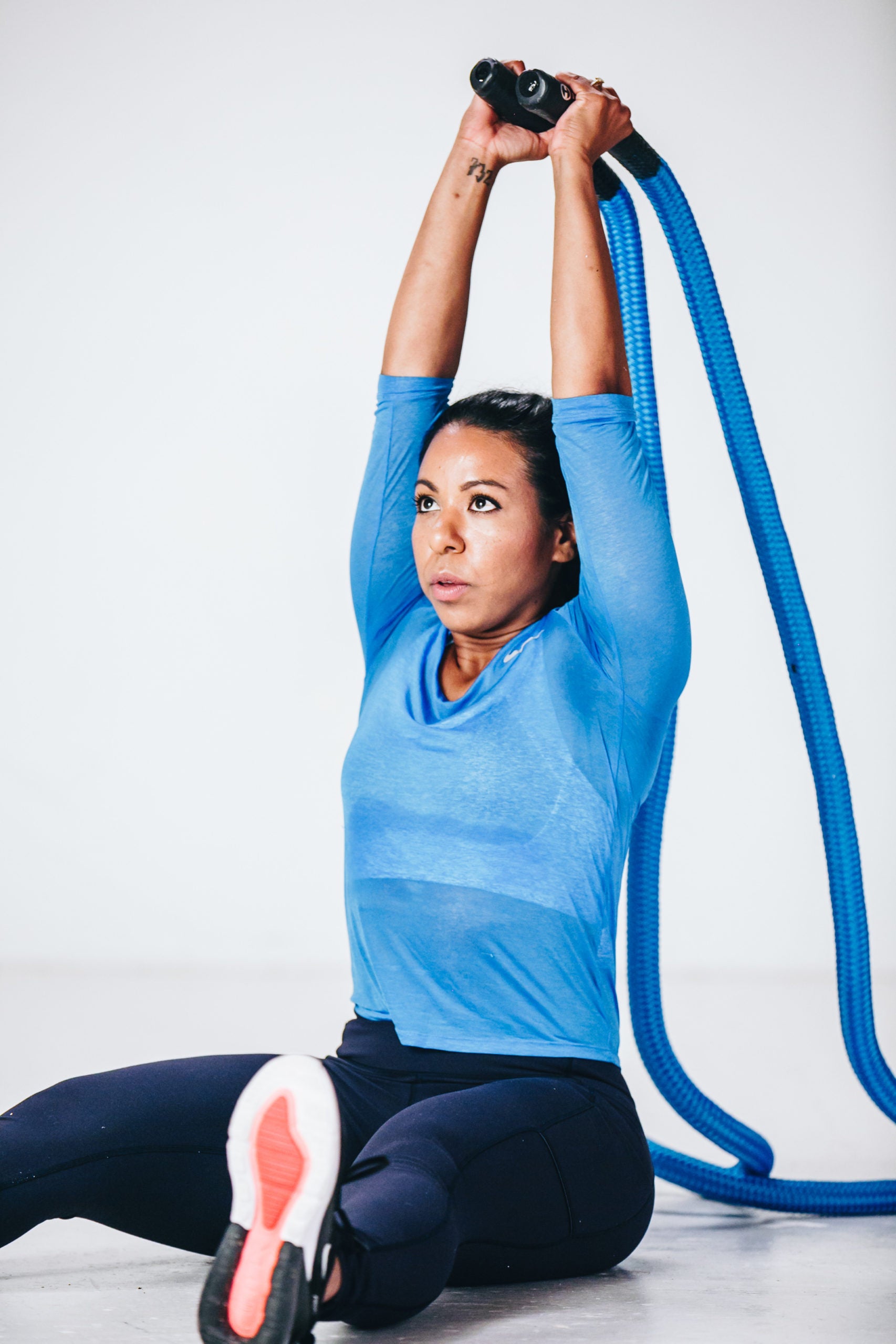 Image of female fitness model seated on floor legs extended and arms extended overhead holding hyper rope alternative battle rope doing overhead tricep extensions battle rope exercise