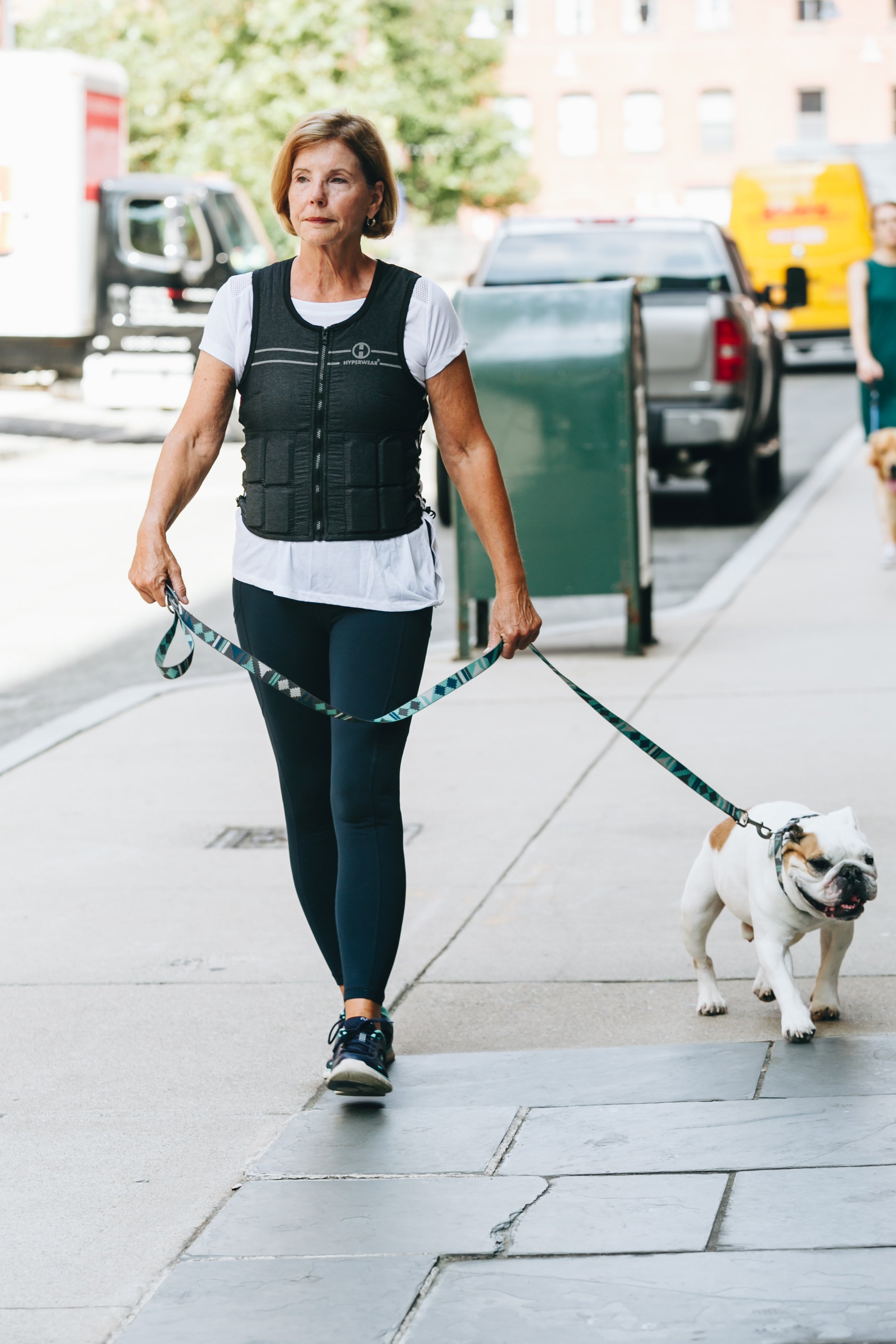 An woman walks her bulldog down a city street while wearing a grey hyper vest fit weighted vest for women to build bone density
