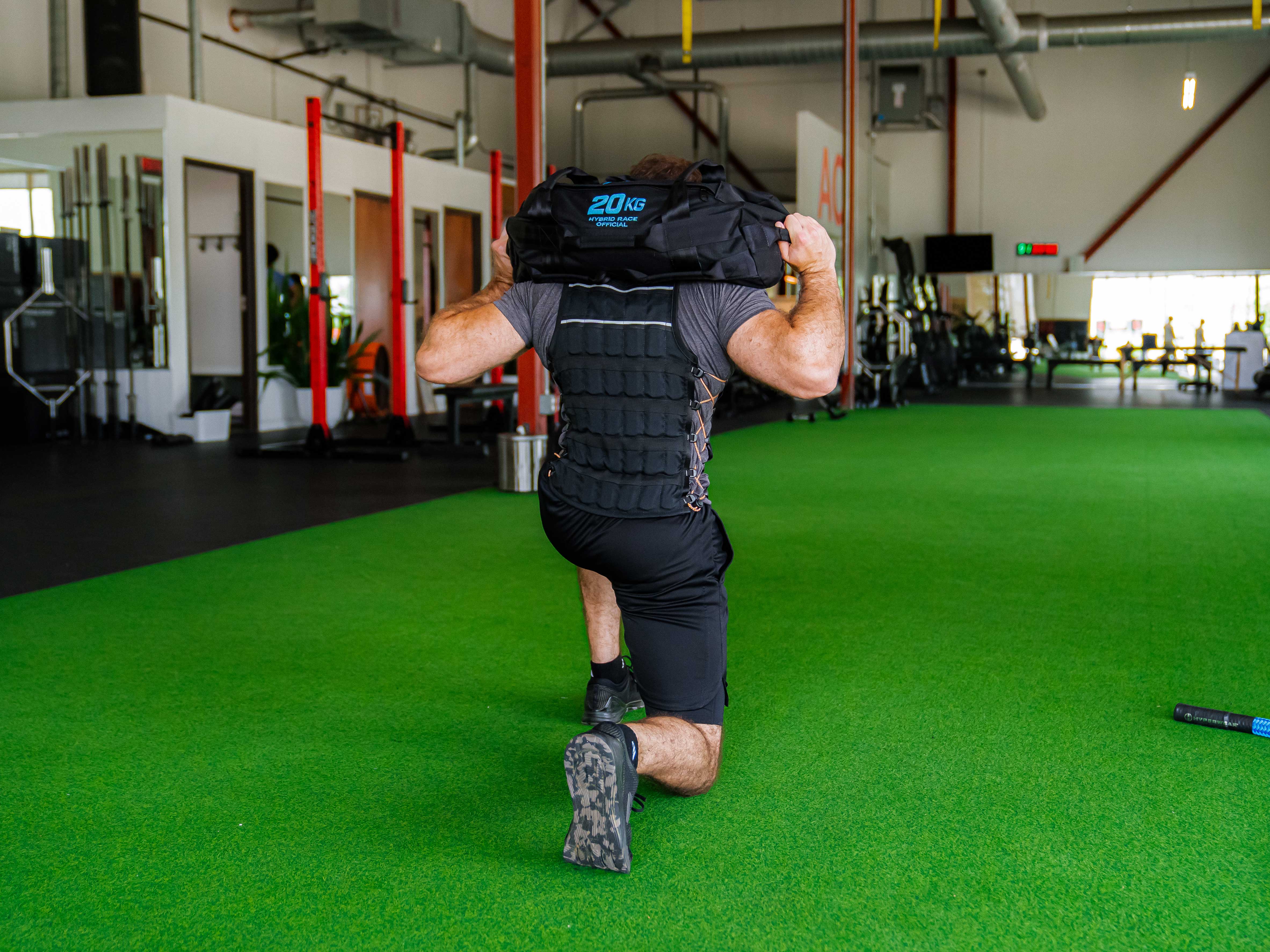 a hybrid racing athlete performs a Hyrox sandbag lunge in a gym