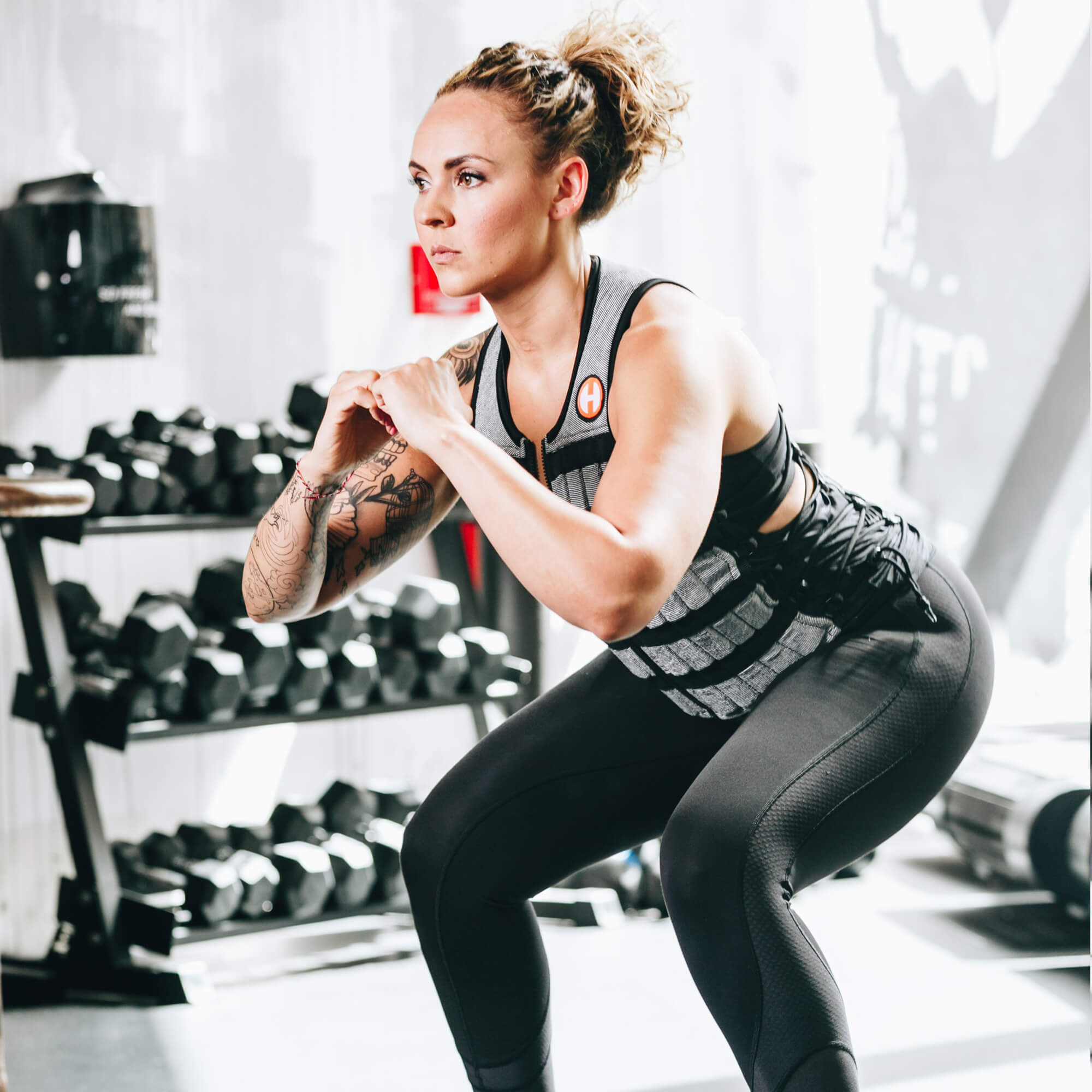 Picture of a woman in a gym in front of a dumbbell rack doing a squat with a hyper vest pro to ad to the bodyweight training benefits