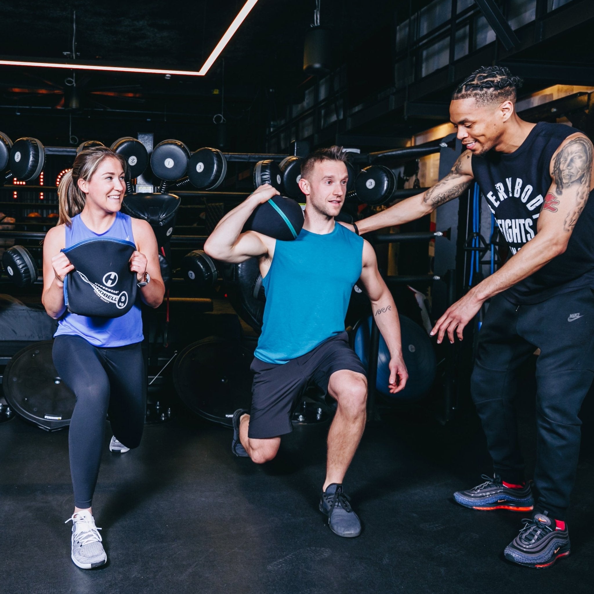 picture inside a health club with two members doing lunges holding a SandBell while a trainer gives instructions